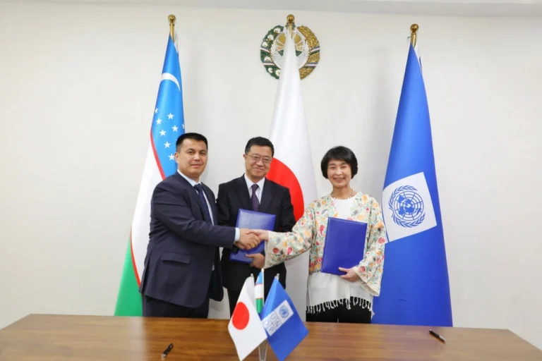 Three officials standing in front of the flags of Uzbekistan, Japan, and the United Nations. A man on the left and a woman on the right are shaking hands while holding blue document folders. A man in a dark suit stands smiling in the center.
